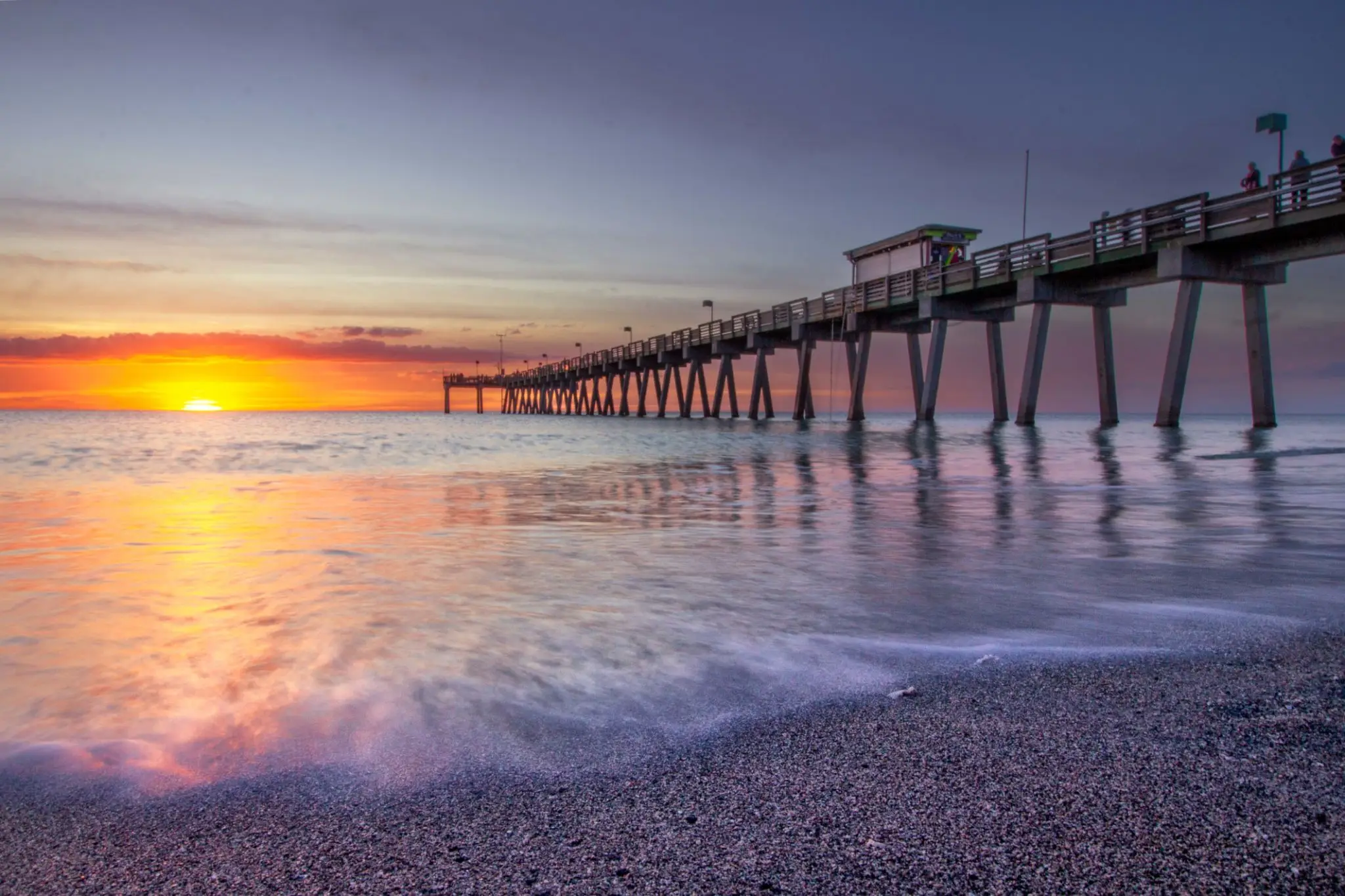Sunset over a calm beach with a long pier extending into the water.