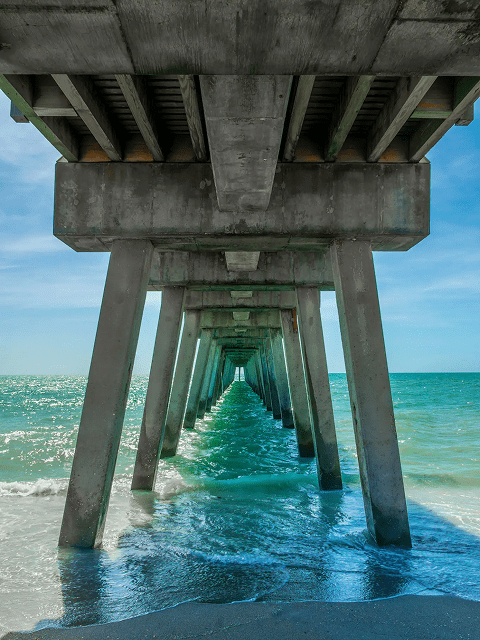 View under a concrete pier stretching into the ocean with clear blue water.